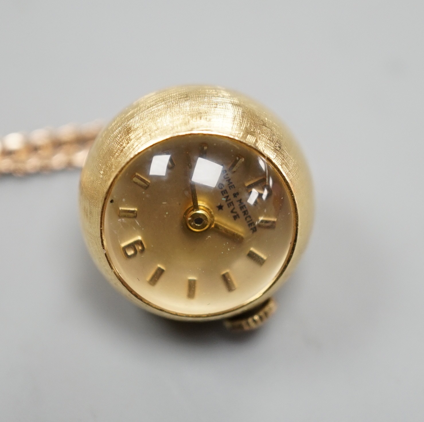 A modern 750 yellow metal Baume & Mercier globe pendant watch, diameter approx. 18mm, on an associated 9ct gold chain, 68cm, chain 6.4 grams.